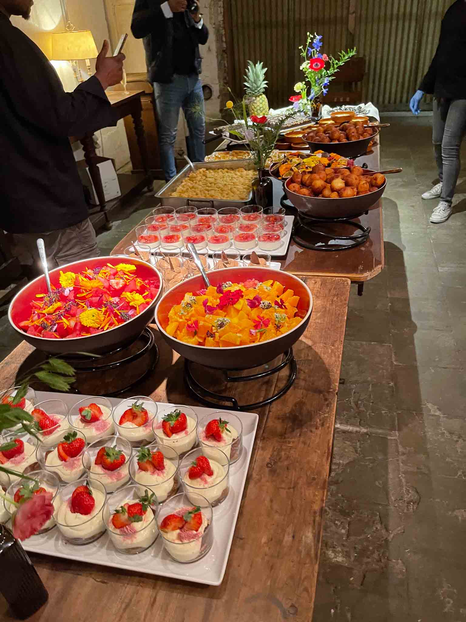 Tafel met etenswaren waaronder tiramisu mousse met aardbeien, chocolade mousse en schalen met vleesbolletjes met bloemenvazen als decoratie.